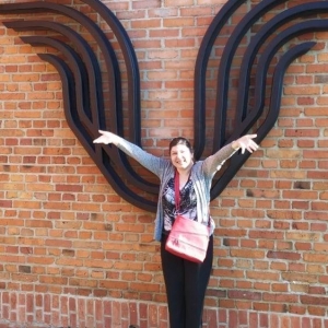 Na zdjęciu widać kobietę stojącą przy murze, na których są zawieszone metalowe skrzydła